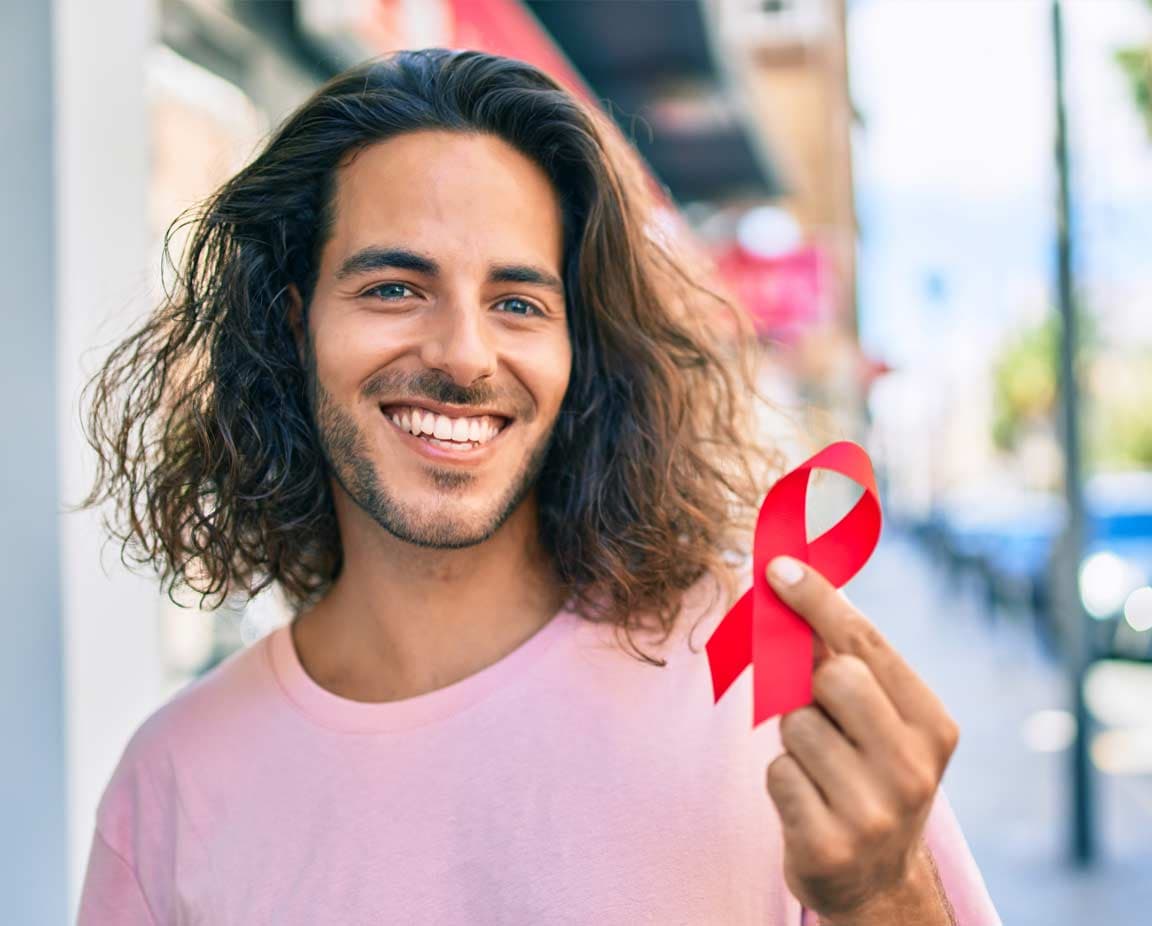 Junger Mann mit langen dunklen Haaren und rosa T-Shirt lächelt in die Kamera und hält eine rote Schleife in der Hand – das weltweite Symbol für Solidarität mit HIV-positiven Menschen.