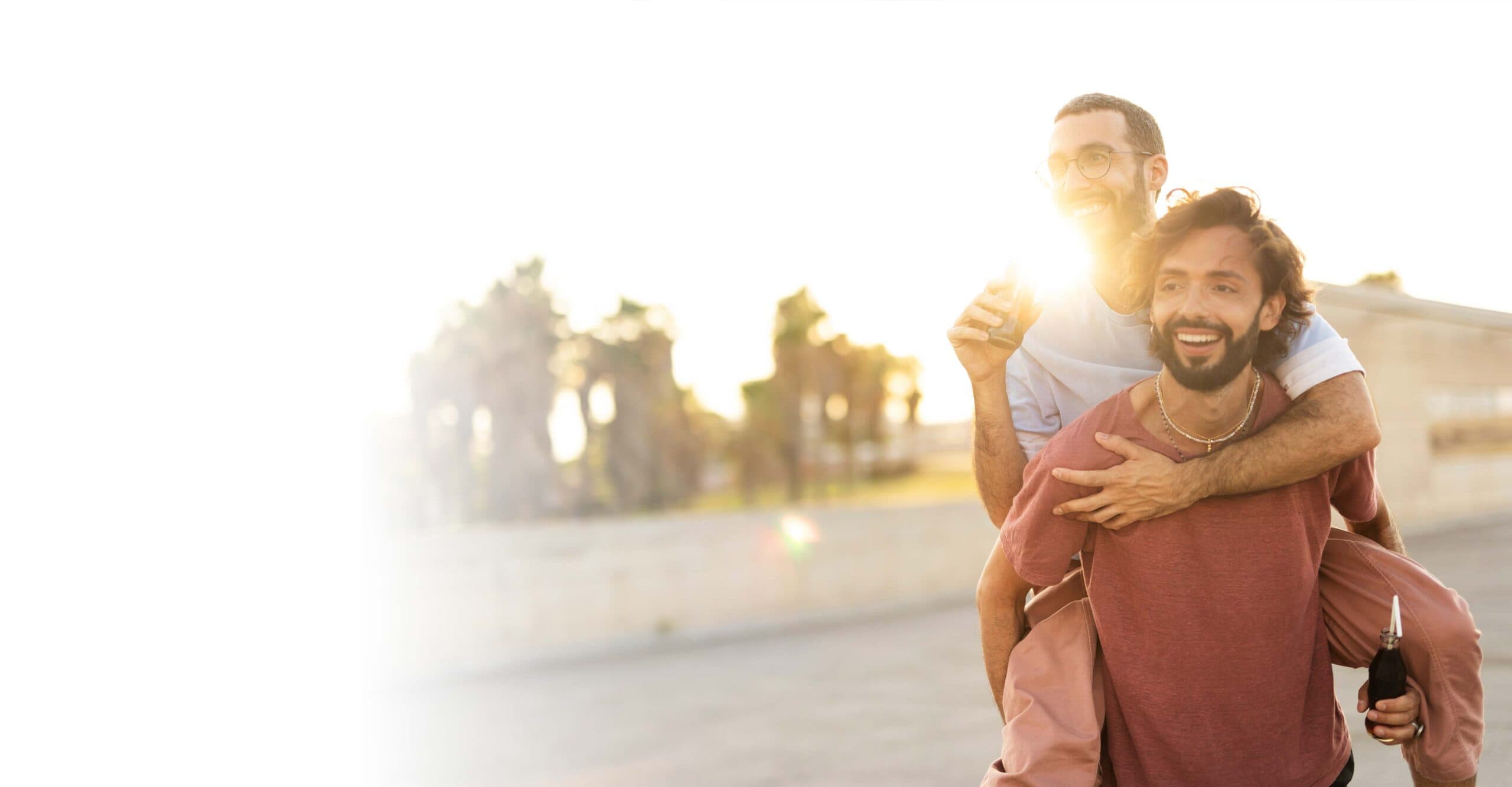 Ein schwules Paar genießt gemeinsam einen sonnigen Moment im Freien. Der eine trägt den anderen lachend auf dem Rücken – Ausdruck von Nähe, Lebensfreude und gegenseitigem Vertrauen.